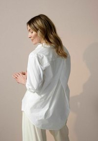 Camicia bianca a maniche lunghe con una vestibilità rilassata, orlo arrotondato e una sottile texture. Sul retro presenta un design incrociato unico. Tessuto morbido e leggero.