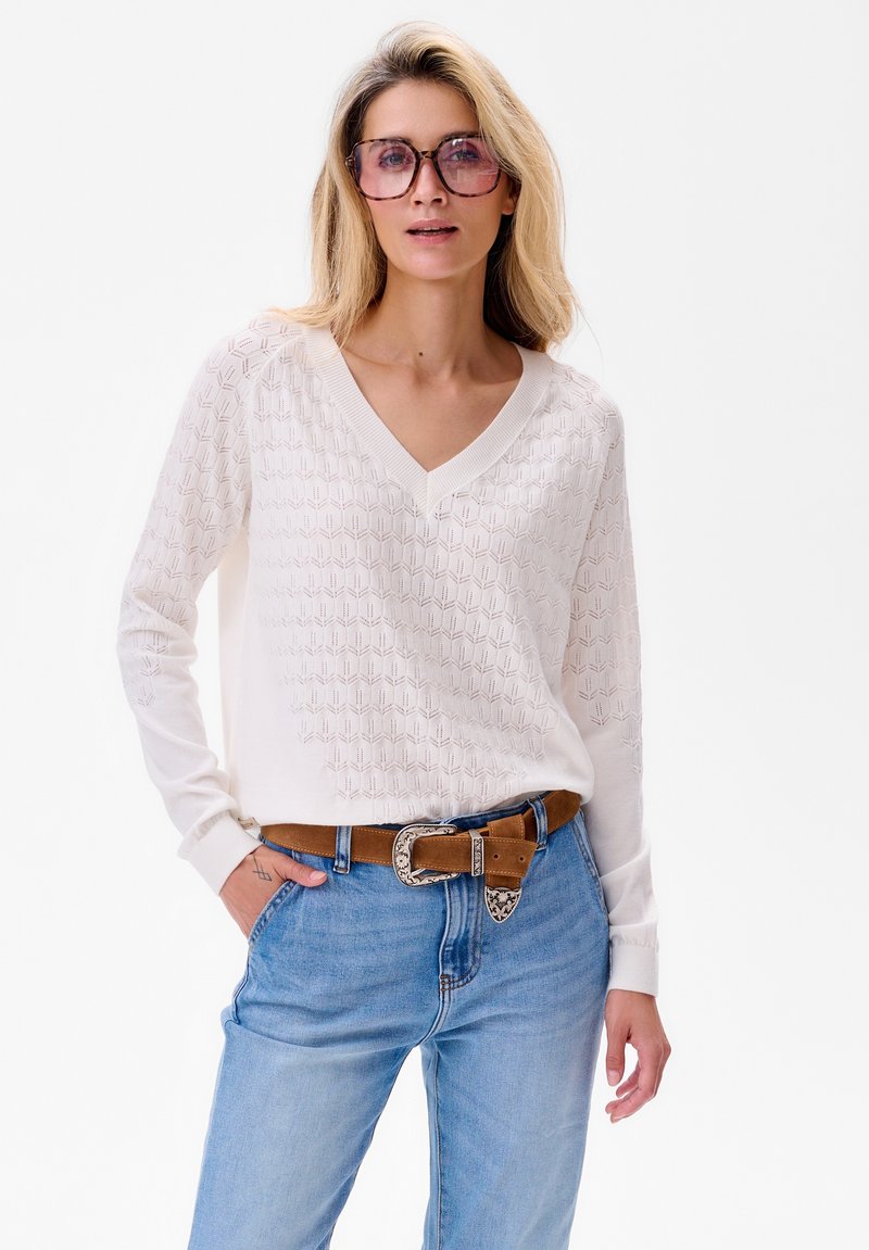 Witte gebreide V-hals trui met een textuur van geometrische patronen. Gecombineerd met blauwe jeans en een bruine riem met een decoratieve gesp.