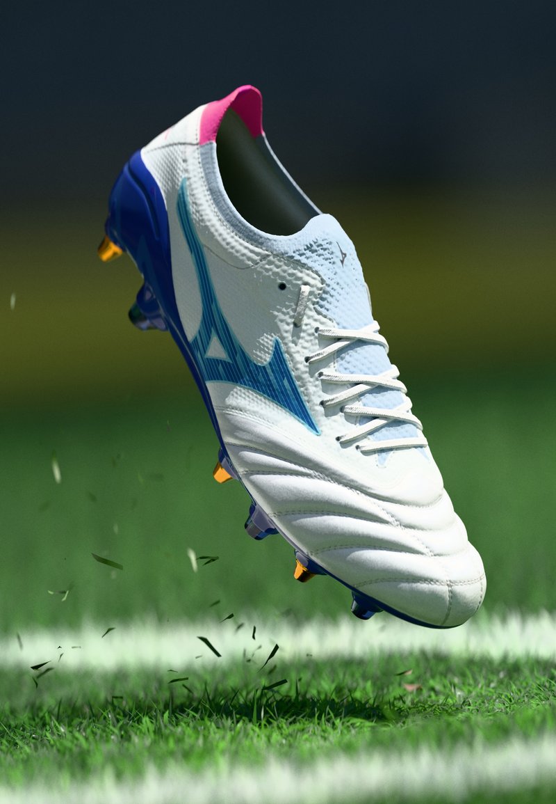Λευκά ποδοσφαιρικά παπούτσια με μπλε λεπτομέρειες και ροζ κολάρο, διαθέτοντας υφή στην επάνω πλευρά και κίτρινους κορυφαίους στύλους, υψωμένα πάνω από το γρασίδι.