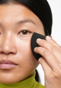 Éponge de maquillage noire tenue contre la peau, appliquant le produit. Gros plan sur une peau lisse, des cheveux foncés et un vêtement vert lumineux en arrière-plan.