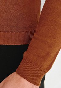 Brązowy sweter z dzianiny z prążkowanymi mankietami i dołem, prezentujący miękką teksturę. Noszony na czarnej odzieży, podkreślający kontrast kolorów.