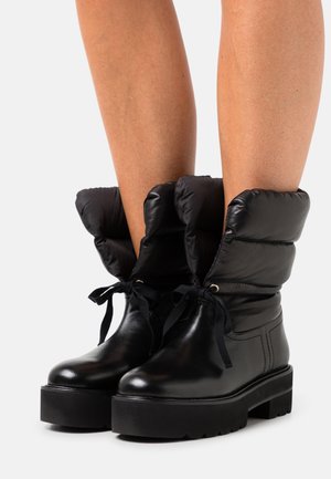 Jalat, joissa on mustat paksupohjaiset platform-saappaat, joissa on pehmustetut, tikatut yläosat ja mustat nauhat valkoista taustaa vasten.