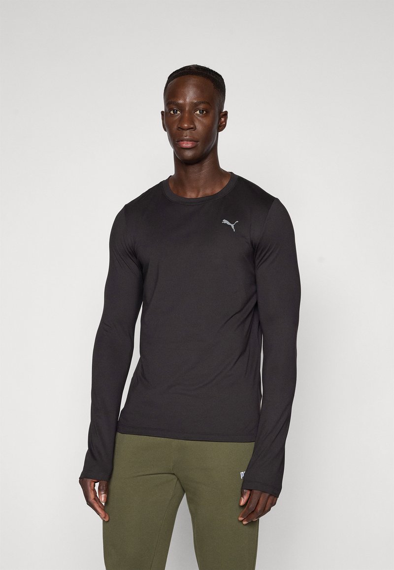 Mann som står iført en tettsittende svart langermet Puma-skjorte og olivengrønne Puma-sweatpants foran en ensfarget lys bakgrunn.