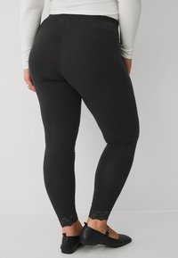 Zwarte leggings met een gladde textuur, voorzien van een kantafwerking bij de enkel en een aansluitend ontwerp. G gedragen met platte zwarte schoenen.