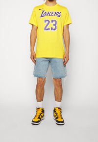 Vuxen person som bär gul Lakers 23 baskettröja, ljusblå slitna jeansshorts, vita strumpor och svart-gula Nike sneakers står upp.