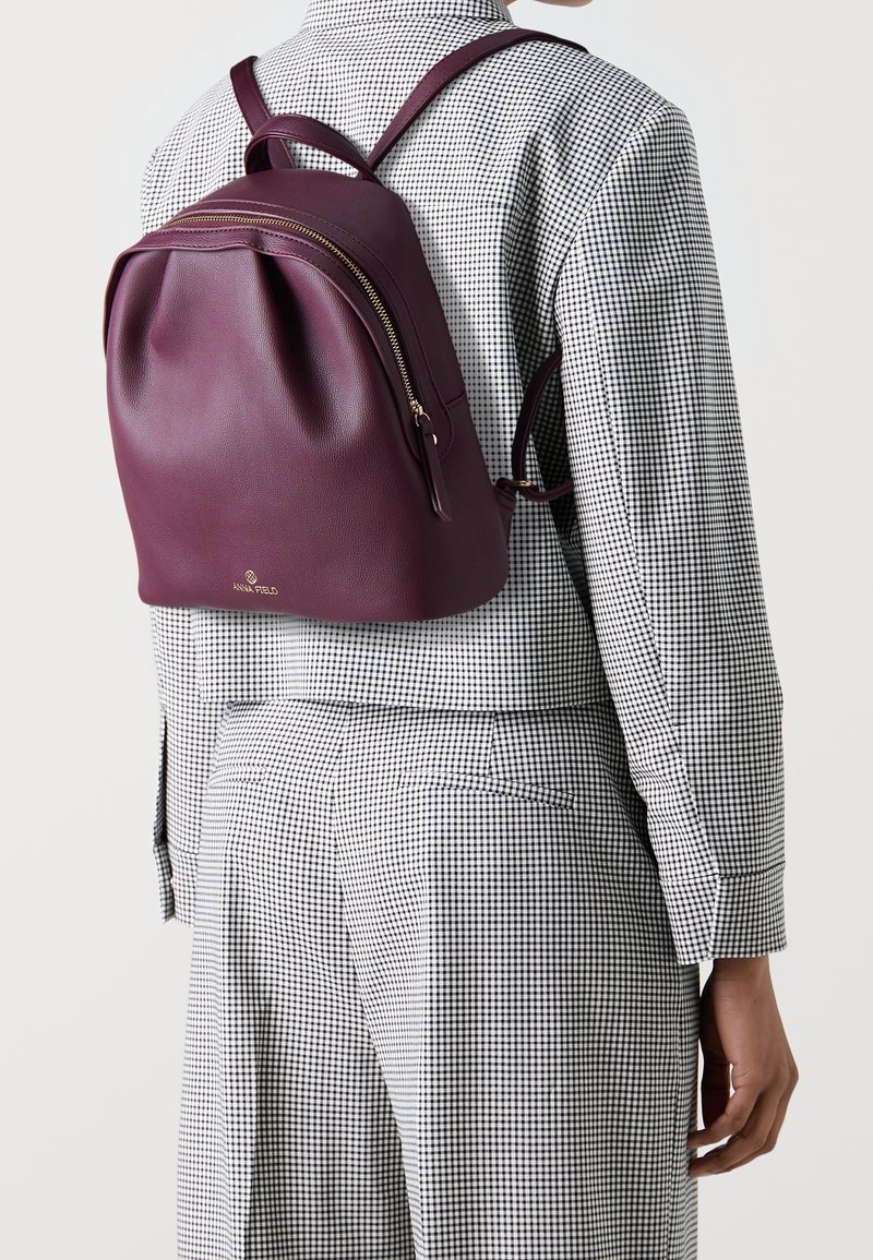Femme portant un sac à dos en cuir violet sur un costume à carreaux noir et blanc, vue de dos et de côté.