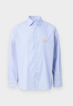 Άνδρας που στέκεται φορώντας ανοιχτό μπλε πουκάμισο με κουμπιά, γκρι παντελόνια φαρδιάς γραμμής και μαύρα και λευκά μοκασίνια με χρυσή αλυσίδα ως διακοσμητικό.