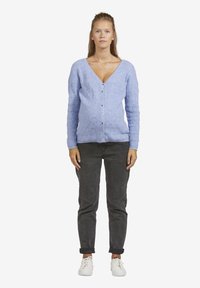 Junge Frau steht mit dem Gesicht nach vorne, trägt einen hellblauen, geknöpften Cardigan, dunkle hochgekrempelte Jeans und weiße Sneaker.