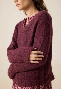 Pull en tricot bordeaux avec un col en V, tissu texturé, larges poignets et ourlet côtelé, superposé à un chemisier à motifs.