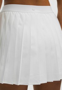 White pleated skirt worn at waist with visible Lululemon logo on smooth waistband against tan skin.