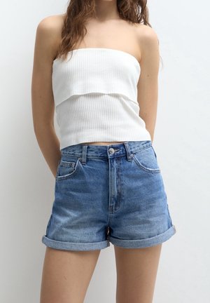 Ung kvinde iført en hvid, skulderfri ribbet top og blå, højttaljede denimshorts, der er rullet op, foran en ensfarvet baggrund.