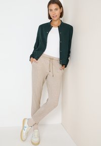 Tmavě zelená, zapínací bunda přes bílou košili; béžové vzorované teplákové kalhoty; bílé tenisky s modrými a hnědými akcenty.