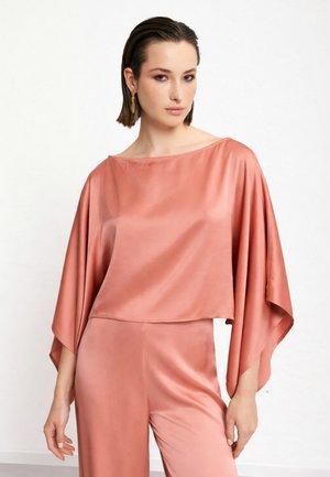 Femme aux cheveux courts et bruns, portant un haut ample en satin rose avec de larges manches et un pantalon assorti, debout devant un fond blanc uni.