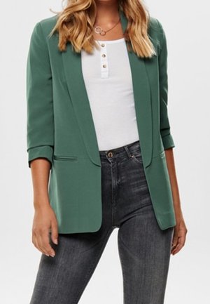 Žena v zeleném blejzru s ohrnutými rukávy, bílém knoflíkovém topu a tmavě šedých džínech s vysokým pasem.