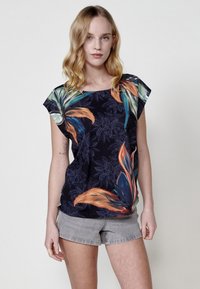 Blouse noire à manches courtes avec des motifs floraux, arborant des feuilles vertes et orange vives, un col rond et une coupe décontractée, assortie à un short gris clair.