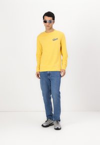 Langärmliges gelbes Hemd mit blauem Logo, blaue Jeans und graue Sportschuhe. Das Modell trägt graue Sonnenbrillen. Sauberer, minimalistischer Hintergrund.