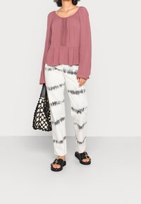 Bluzka peplum w kolorze przybrudzonego różu, z długimi rękawami i misternymi detalami. Spodnie w kolorze białym z wzorem tie-dye i czarnymi akcentami; czarne sandały.
