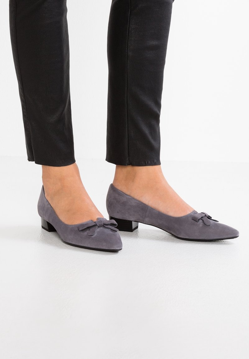 Chaussures basses en daim gris avec un bout pointu et un nœud décoratif à l'avant. Texture lisse et design simple, associées à un pantalon noir.