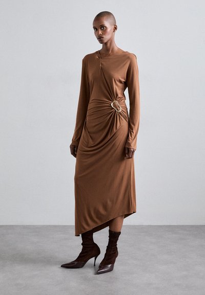 Vestido marrón de manga larga con detalle drapeado y un acento de anillo dorado en la cintura. Estilizado con botas de tacón puntiagudas marrones. Textura suave.