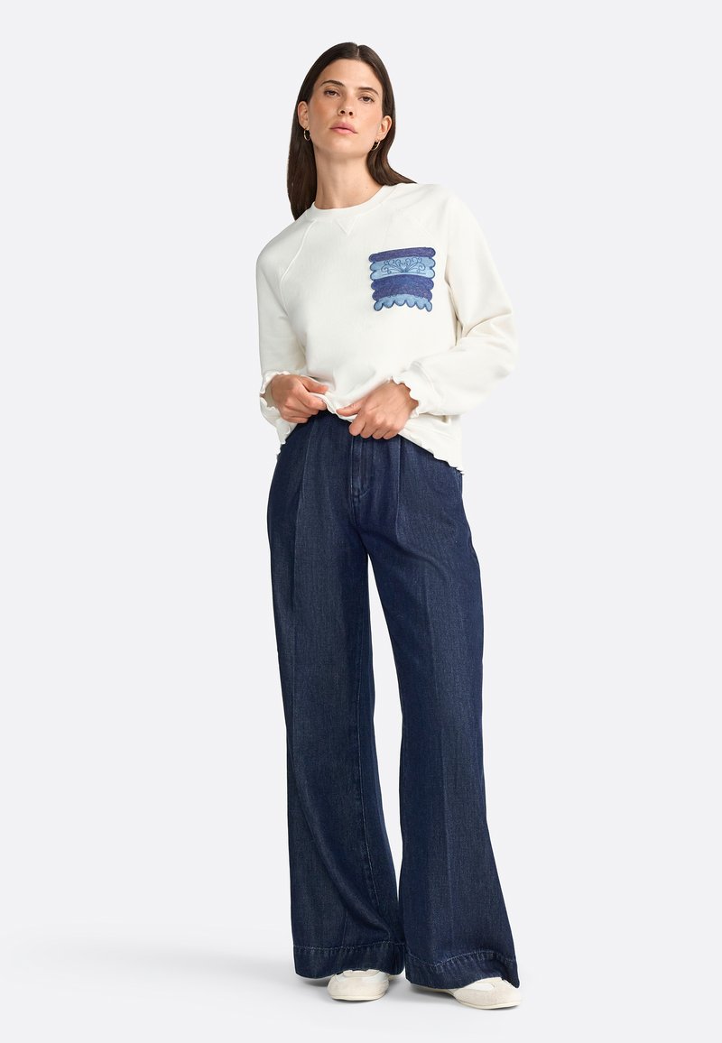 Sudadera blanca de manga larga con un bolsillo azul con volantes, combinada con jeans de pierna ancha de mezclilla oscura y zapatos de color claro.