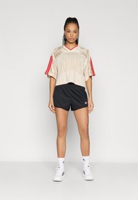 Jersey corto beige con rayas rojas en las mangas, con textura de malla y gran logo impreso, combinado con pantalones cortos negros y zapatillas blancas.