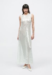 Hosszú, ujjatlan ruha, áttetsző, ezüstszínű, tükröződő flitterekből, egyenes sziluettel és földig érő fazonnal.