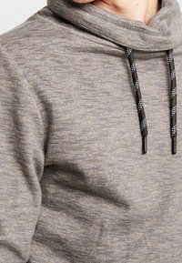 Grå pullover med cowl-neck, som har et tekstureret stof og sorte snore med hvide accenter. Glat, blød struktur synlig.