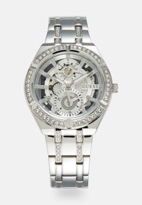 Orologio in metallo argentato con quadrante scheletrato trasparente, cristalli decorativi sulla lunetta e sul bracciale, con tre sotto-quadranti e logo 'GUESS'.