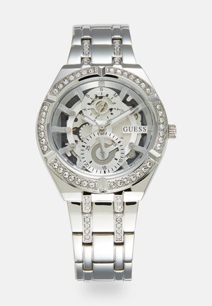 Silver metal watch with a transparent skeleton dial, decorative crystals on the bezel and bracelet, featuring three subdials and 'GUESS' logo.