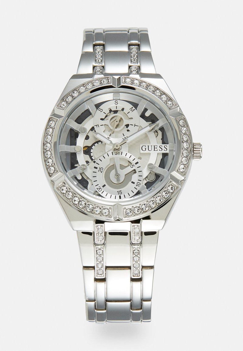 Orologio in metallo argentato con quadrante scheletrato trasparente, cristalli decorativi sulla lunetta e sul bracciale, con tre sotto-quadranti e logo 'GUESS'.