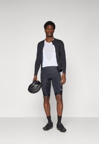 Veste de cyclisme noire sur un haut en maille blanc, shorts noirs avec rembourrage, chaussures de cyclisme noires, tenant un casque noir.