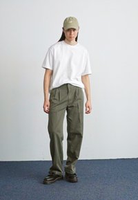 Bijela oversized majica uparena s maslinasto zelenim hlačama visokog struka i crnim chunky cipelama. Outfit uključuje svijetlozeleni kapu i opušteni kroj.
