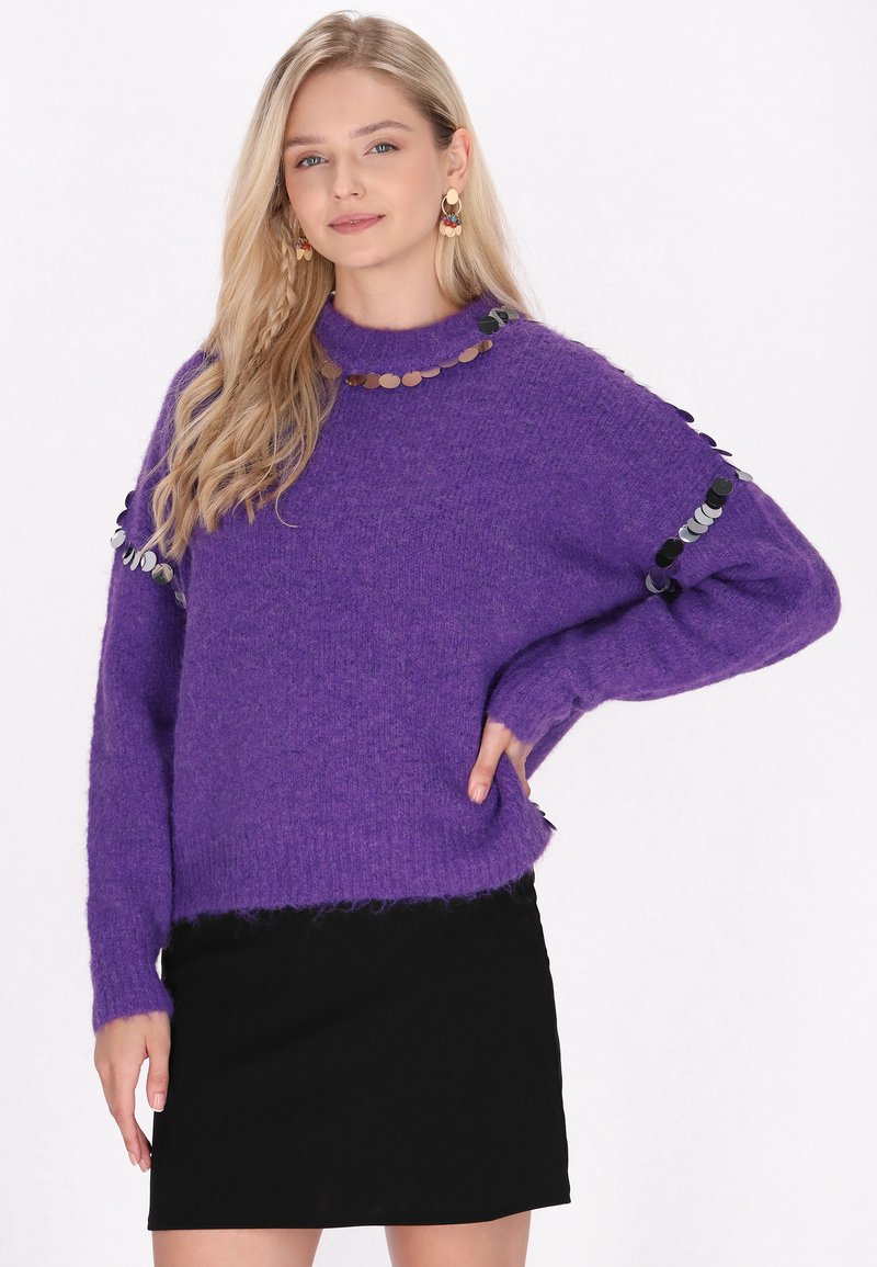 Lila, strukturiertes Strickpullover mit hohem Kragen, verziert mit runden Pailletten entlang des Kragens und der Ärmel. Getragen über einem schwarzen Rock.