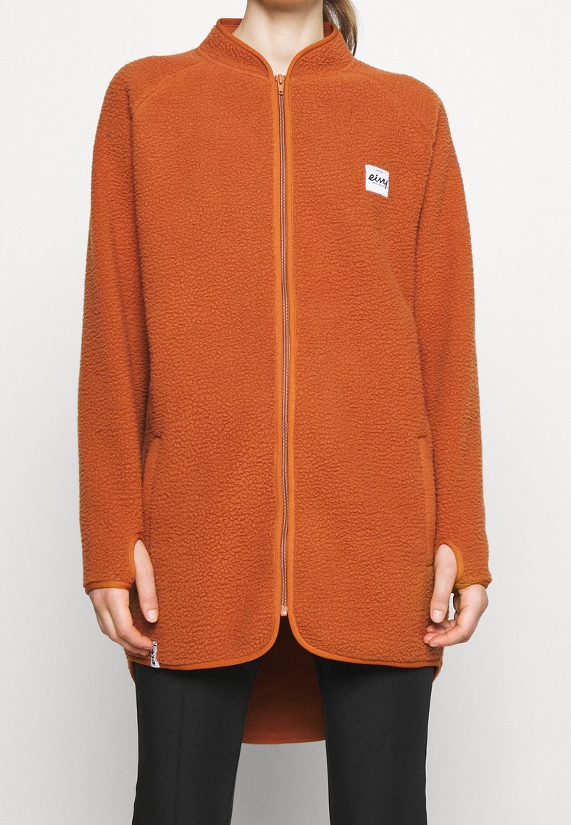 Femme portant une longue veste polaire orange à fermeture éclair avec des trous pour les pouces, associée à un pantalon noir, debout devant un fond uni.