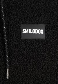 Svart fleece tyg med en texturerad yta, med en vävd dragsko och en rektangulär svart etikett med vit text "SMILODOX."