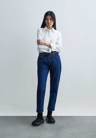 Žena stoji s prekriženim rukama, odjevena u bijelu košulju na dugih rukava, plave traperice visokog struka s crnim remenom i crne platforme.