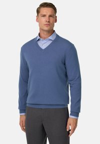 Mėlynas V formos megztinis iš trikotažo su raukiniais ant rankovių ir apatinės ilgos dalies, uždėtas ant šviesiai mėlyno dryžuoto marškinėlių, derinamas su pilkomis kelnėmis.