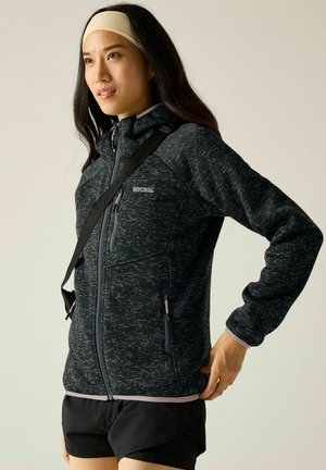 Femme portant une veste zippée gris foncé, un short noir, un bandeau beige, et une sangle de sac en bandoulière sur l'épaule, regardant sur le côté.