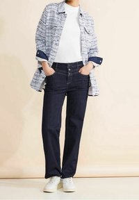 Vit kortärmad skjorta under en blå och vit mönstrad skjorta, kombinerat med mörkblå jeans och vita sneakers. Jeans har knäppning.