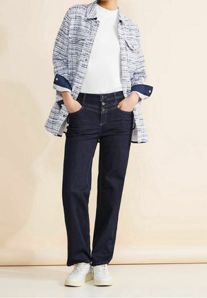 Camicia bianca a maniche corte sotto una camicia blu e bianca a fantasia, abbinata a jeans blu scuro e sneakers bianche. I jeans hanno una chiusura con bottoni.