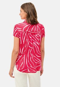 Blusa rosa a maniche corte con motivi di foglie bianche, vestibilità rilassata, dettaglio della piega sul retro e orlo leggermente curvo, abbinata a pantaloni chiari.
