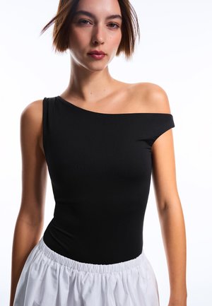 Vrouw met kort bruin haar, die een zwart top met één schouder en een witte geplooide rok draagt, tegen een witte achtergrond.