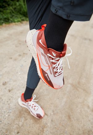 Personne portant des chaussures de trail Gore-Tex rouges et beiges avec des lacets blancs et des chaussettes noires sur un sentier en terre.