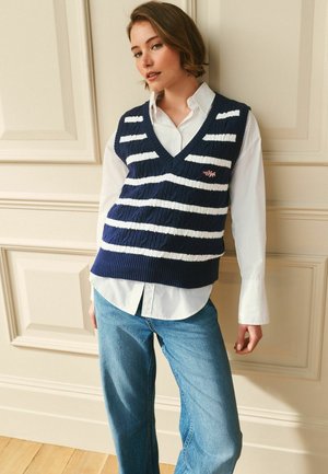 Junge Frau lehnt an einer cremefarbenen Wand, trägt ein weißes Hemd, eine marineblaue und weiß gestreifte Strickweste und blaue Jeans.