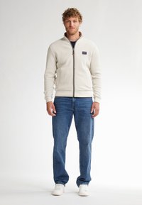Světlobéžová zipová bunda z texturované pleteniny, s stojáčkem, žebrovanými manžetami a pasem, nošená přes námořnickou košili a modré džíny.