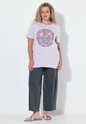 Frau mit lockigem blondem Haar trägt ein lavendelfarbenes Shirt mit einem pinken Leoparden-Print-Smile-Gesicht und graue Leoparden-Print-Hose mit weitem Bein.