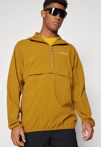Ľahká pulóverová bunda horčicovej farby, s polovičným zipsom na prednej strane, nastaviteľnou kapucňou a veľkou prednou kapsou so elastickými manžetami.