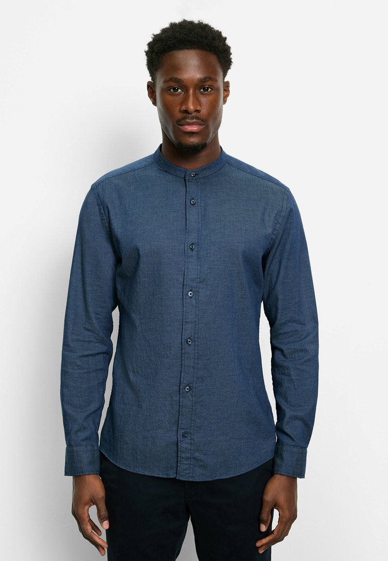 Junger Mann, der frontal steht, trägt ein knopfgeknöpftes, dunkelblaues Hemd mit Stehkragen und schwarze Hosen vor einem schlichten weißen Hintergrund.