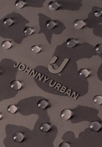 Wassertropfen auf dunkelgrauem Camouflage-Stoff mit erhabenem "Johnny Urban"-Logo und stilisiertem "J"-Emblem.