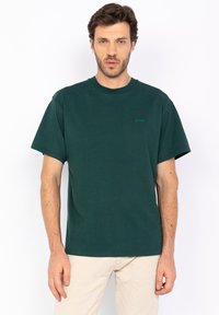 T-shirt in cotone verde scuro, con maniche corte, collo a giro e un piccolo logo verde sul lato sinistro del petto. Texture liscia, vestibilità rilassata.